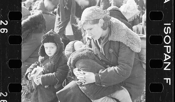 A mother cradles a child while another stands close by. They wear winter clothes of the 1940s and are amidst others waiting.