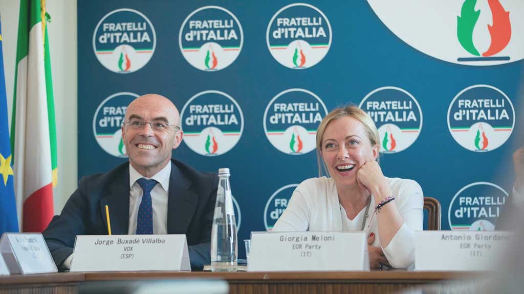 two politician site at a press conference desk and laugh, behind them is a backdrop of the political party's logo.