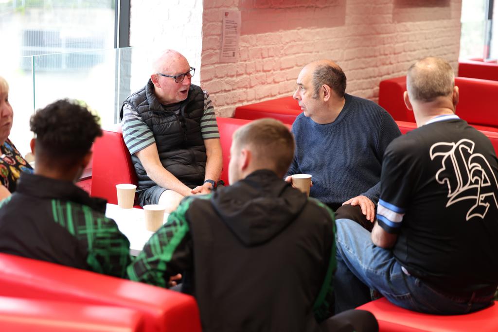 A group of old and young men sit on red sofas, listening to one of themselves.