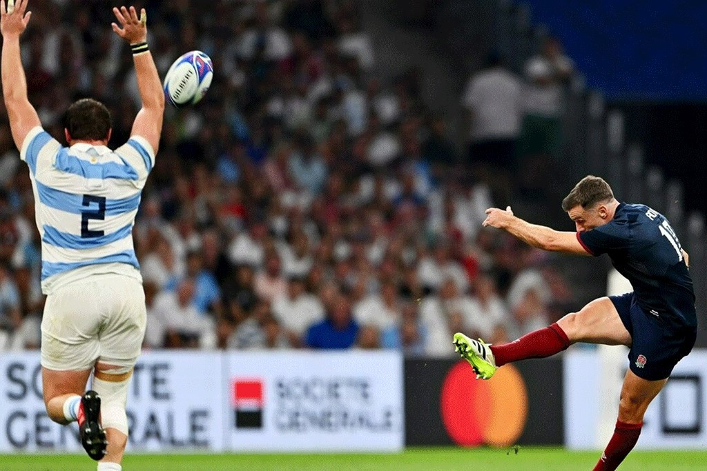 A rugby ball sails towards a player in a striped jersey from the foot of a kicker who has a leg and an arm extended out.