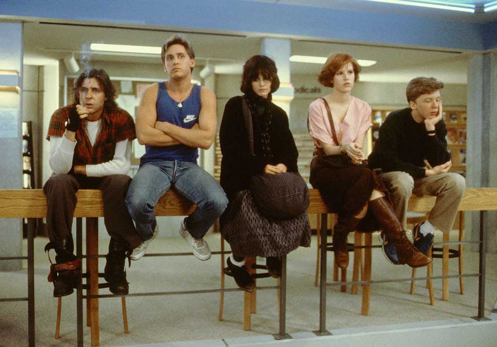 Five glum students perch on stools in school room.