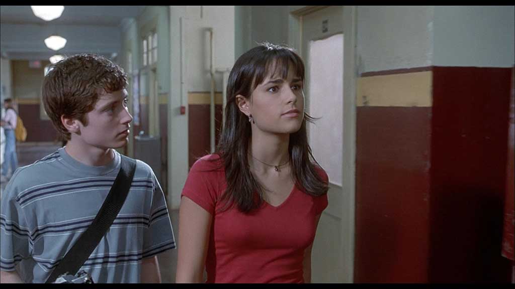 Two students walk down a school corridor, one looking away.