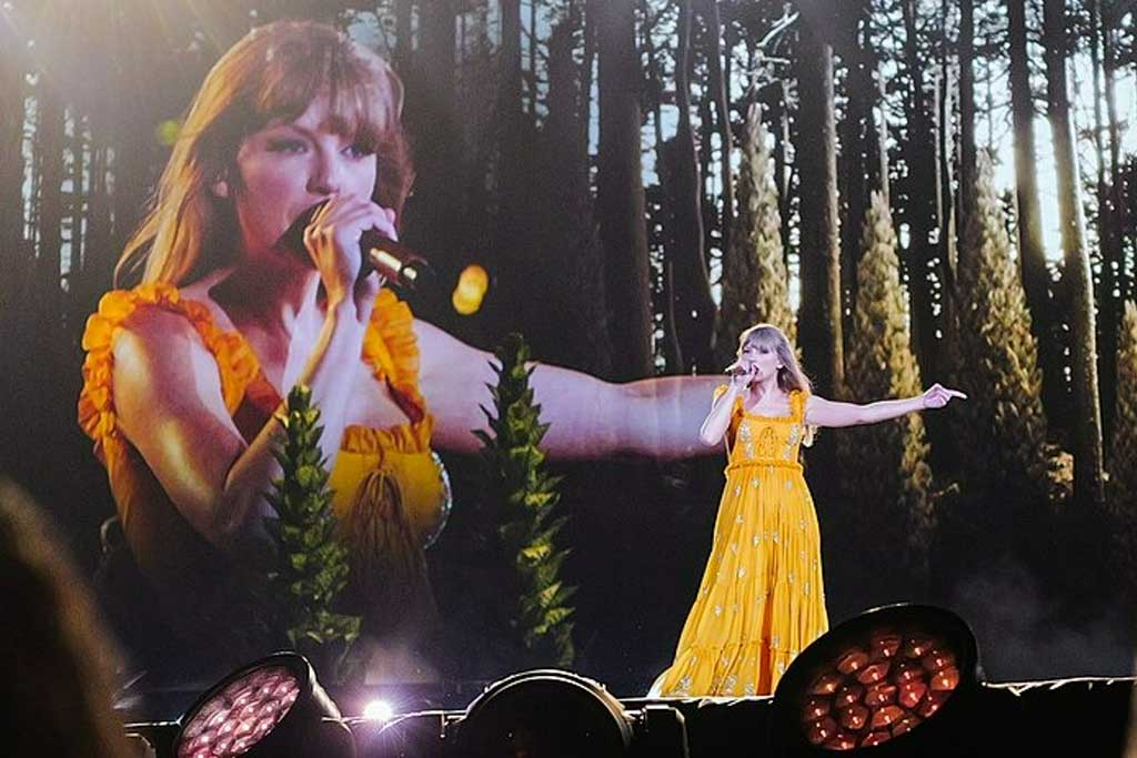 Taylor Swift wears a long yellow dress and signs with an outstretched arm against a backdrop of woods and a close up her singing beside it.