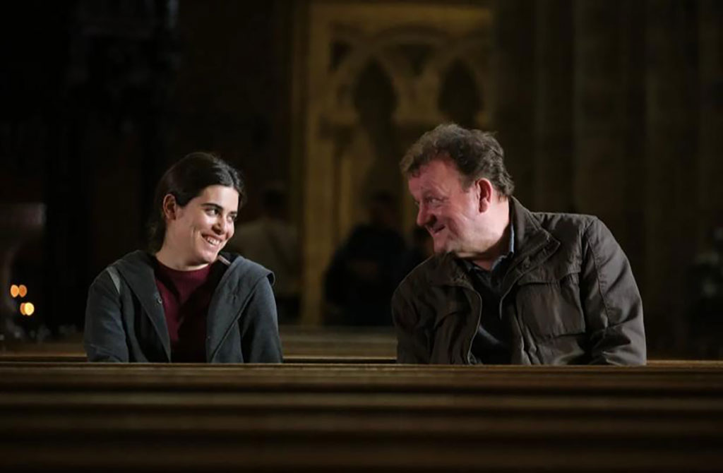 A man and a woman sit in a cathedral pew and incline their heads towards each other.