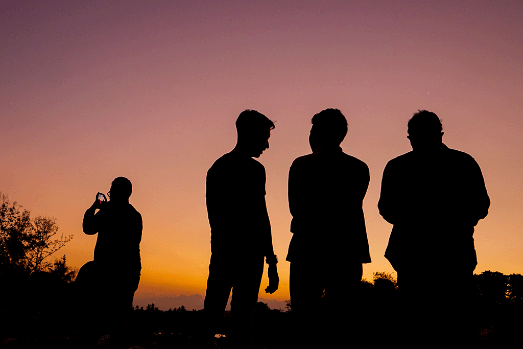 Four men stand silhoutted against a sunset, One stands apart on their phone.