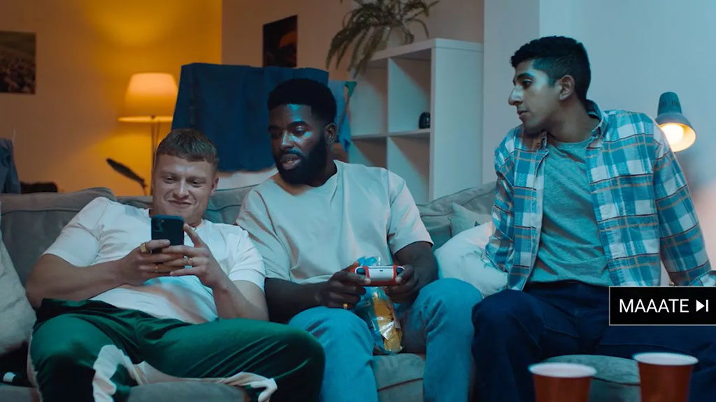 Three young men sit on a couch. One is leering at a phone while the others look on hesitantly