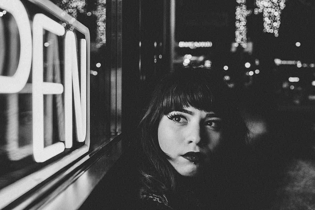 A black and white close up of a women in a street at night, turning to look around at a neion 'open' sign.