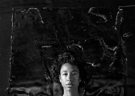 A black and white portrait of a young woman's head against a dark textured background.