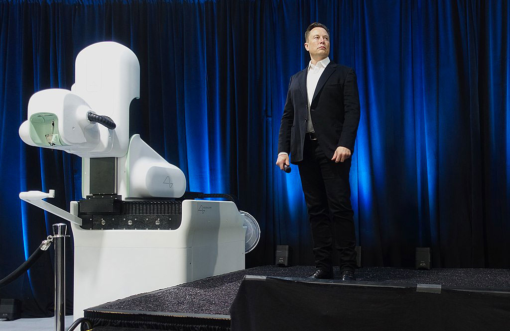 Elon Musk, wearing a dark suit, stands on a stage to a white robotic looking surgical robot.