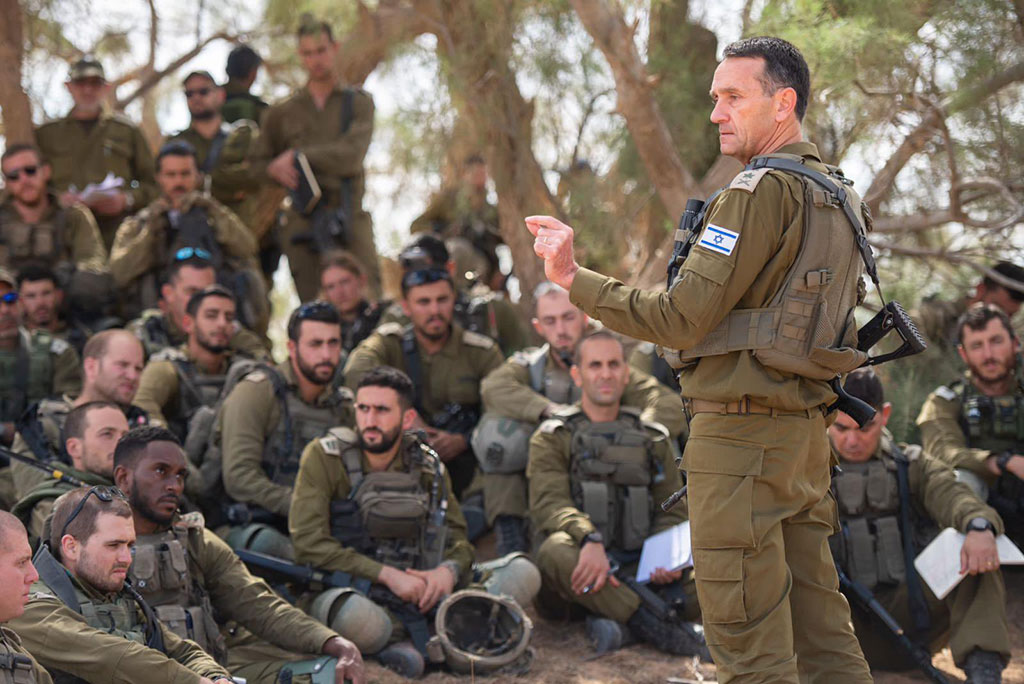 An army general in green fatigues stands and address a group of soldiers sitting, squatting and standing around him