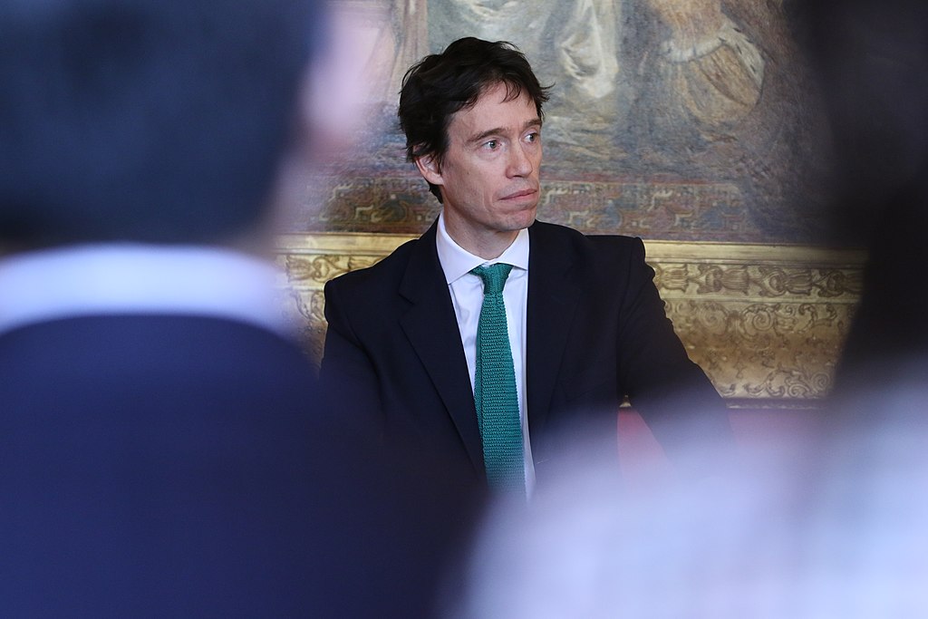 A suited politician stands looking pensive, framed by two out of focus audience members.