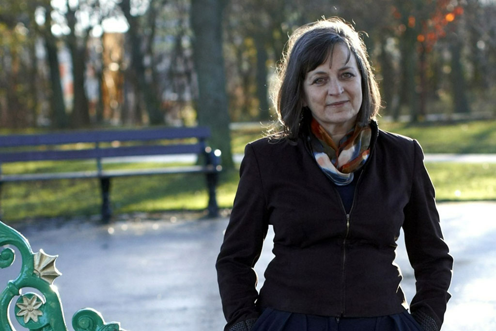 A woman stands in an autumnal-looking park, with her hands in her pockets