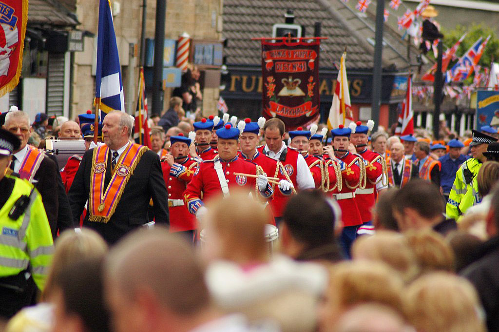 Across the heads of a roadside crowd, men wearing orange sashes and military band uniforms march along.