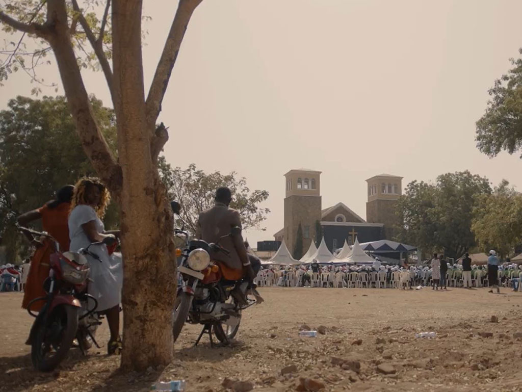 Outside a church a congregation waits seated while an onlooker rests on a motorcycle by a tree.