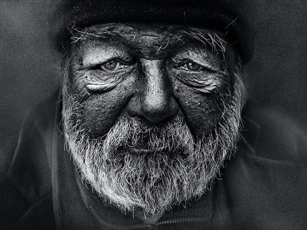 A black and white close up of the weather-beaten and wrinkled face and beard of a homeless man.