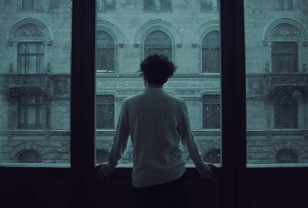 A person stand looking out a wide set of windows covered in rain.