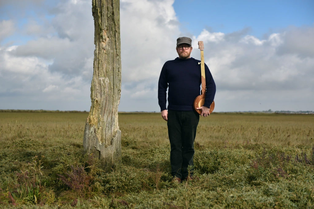 Standing on a salt marsh beside a wooden pillar, a man holds a banjo upright like a rifle.