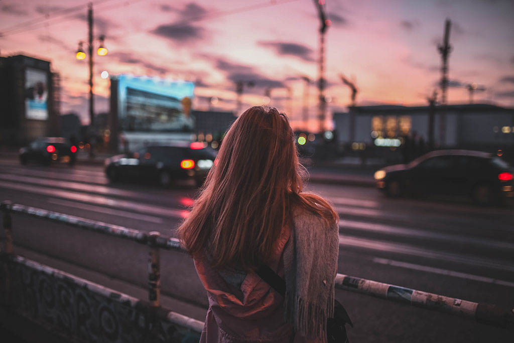 A woman stand beside a busy road with her back to the camera. There is a red sky behind her.