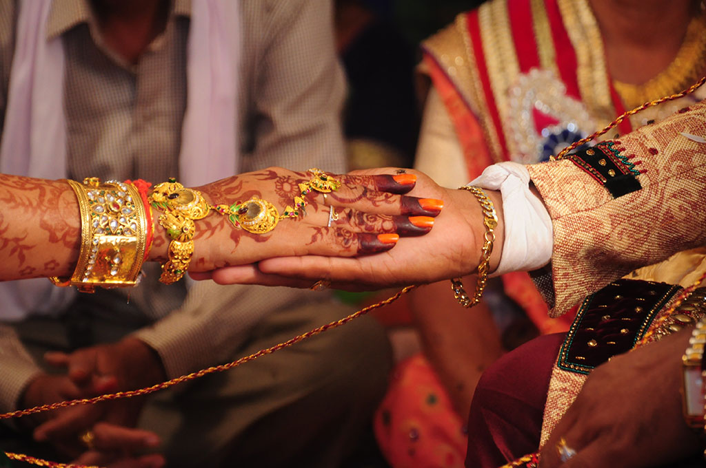 A close-up of a bride groom holding the brides hand. Her hand is henna tattooed and bears gold rings and bracelets. 