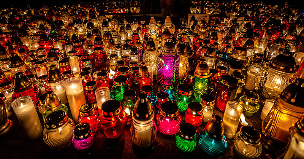 Dozens of candles in cloured jars and holders litter the ground of a cemetry.