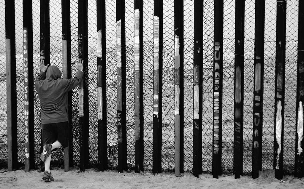 A person holds the vertical tall steel bars of a border fence.