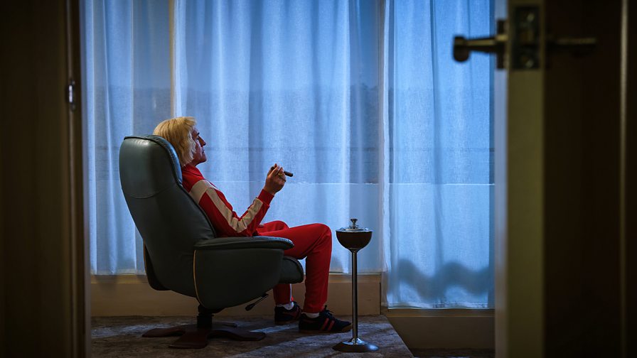 A man in a tracksuit sits in an arm chair smoking a cigar while looking towards a curtained window.