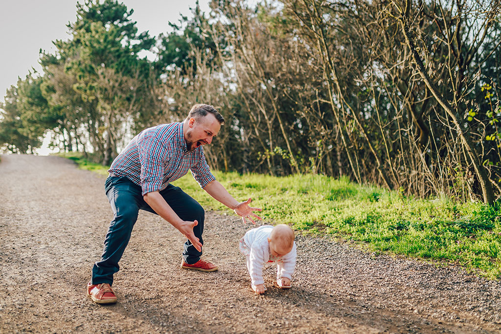 A dad hovers with open arms ready to catch a baby taking first steps