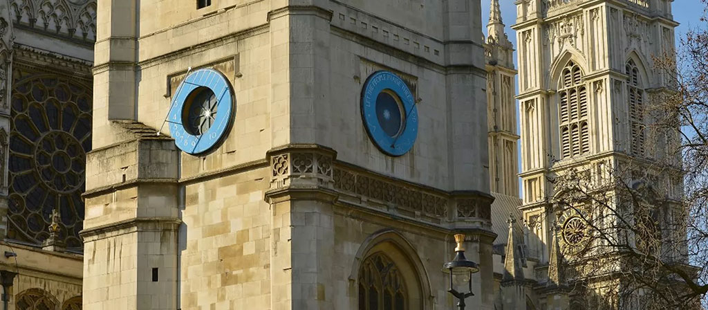 A close up of a blue sun dial on a church clock tower, with an abbey's windows behind it.