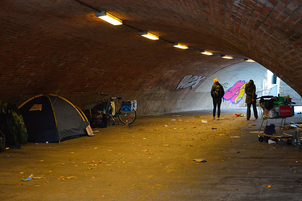 In an underpass a pedestrian passes and look at the tent of a homeless person.