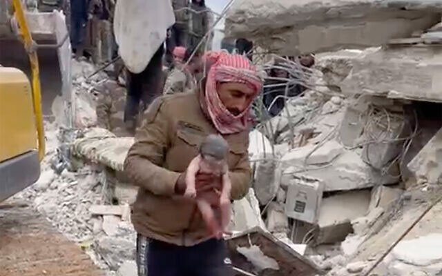 In front of a collapsed building, a rescuer carries a new born baby by the arms.