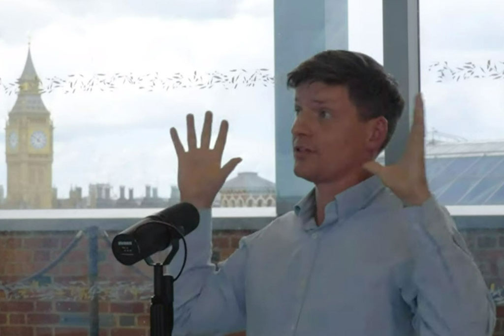 A man sits at a table recording a podcast and holds his hands up for emphasis.