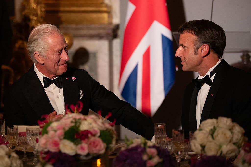 King Charles in a black dinner suit talks and gestures to President Macron who is similarly dressed.