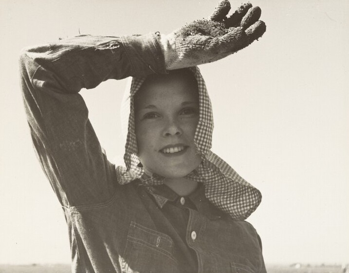 A black and white picture shows a farm worker wearing a headscarf and holding her arm over her head for shade.