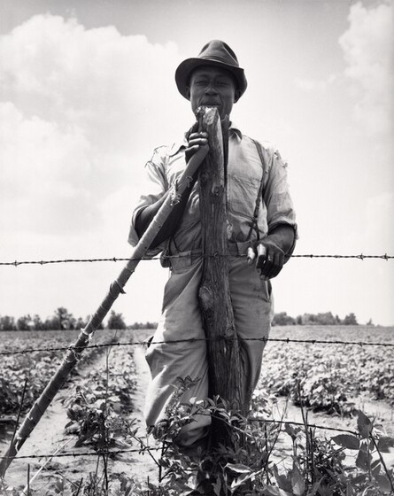 A  farmer stands and leans against an implement he hold with one hand.