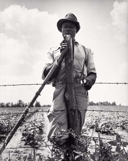 A farmer stands and leans against an implement he hold with one hand.