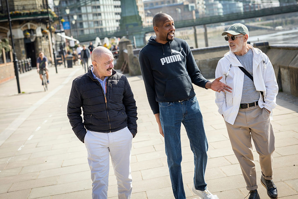 Three men walk down a path, the middle one talking and gesturing while the others listen.