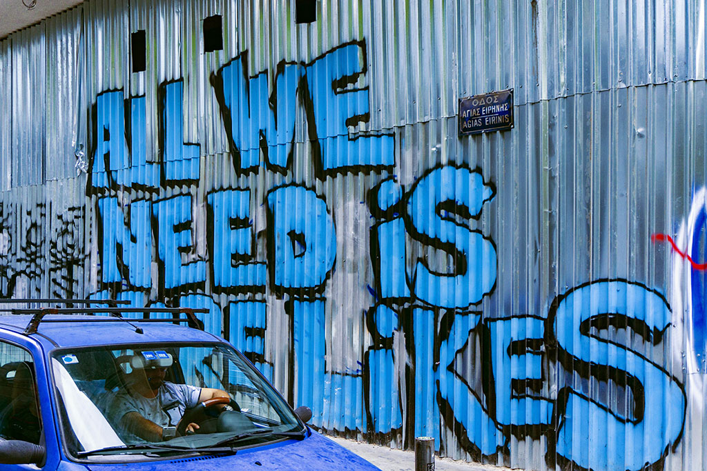 A corrugated sheet iron wall graffited in large blue letters that say 'All we need is more likes.'