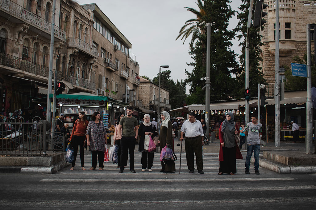 A line of people, some old, some young, wait to cross a road.