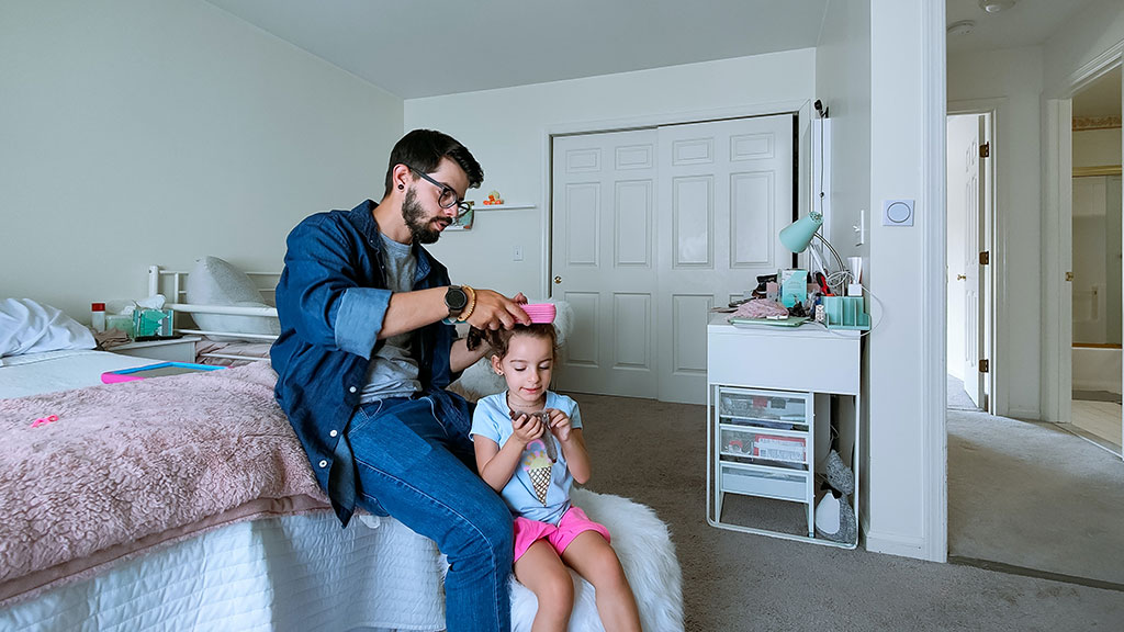 A father sits on a bed and fixes the hair of his daughter standing in front of him