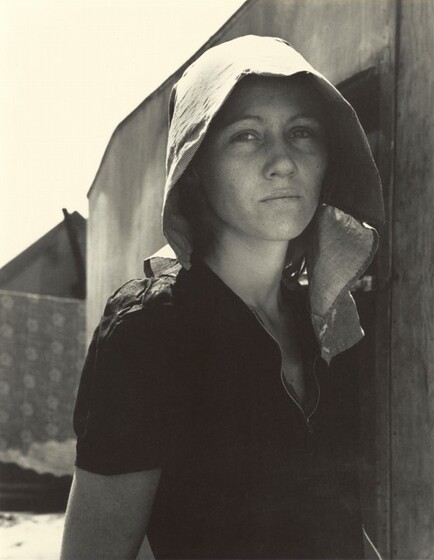 A young woman wearing a sun hat leans against a wall seeking shade.