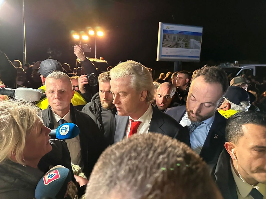 A politician in a suit stands amid a scrum of reporters holding microphones