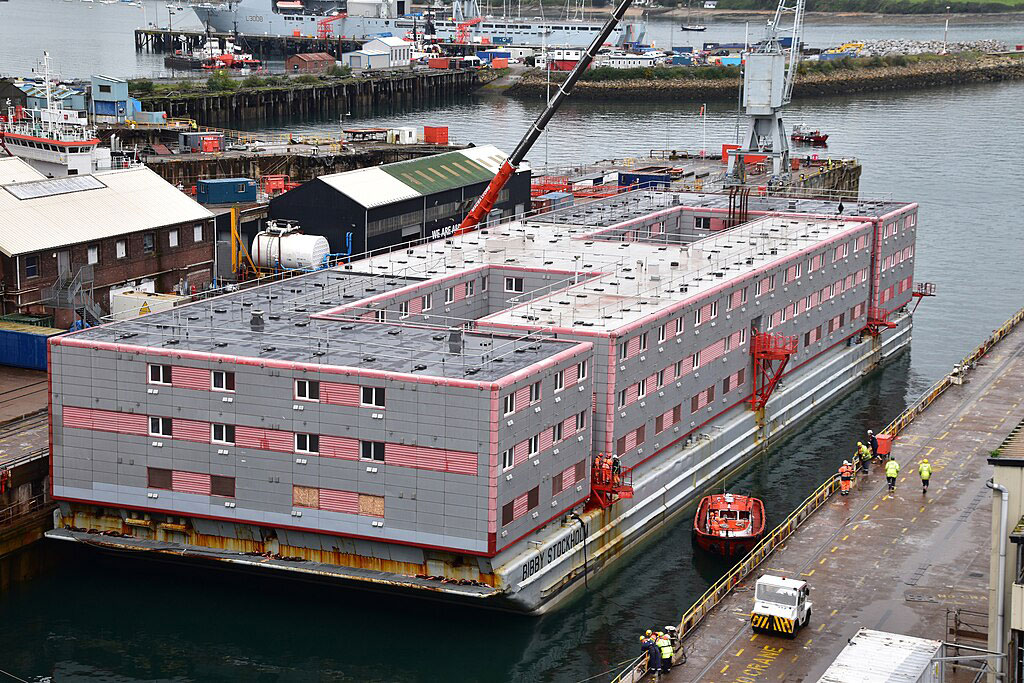 A grey multi-story accommodation barge floats beside a dock.