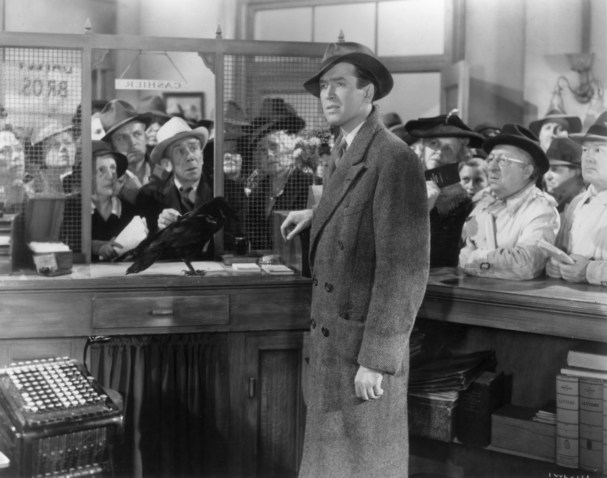 A man stands one side of a bank counter while others, on the other side, look hopefully at him.