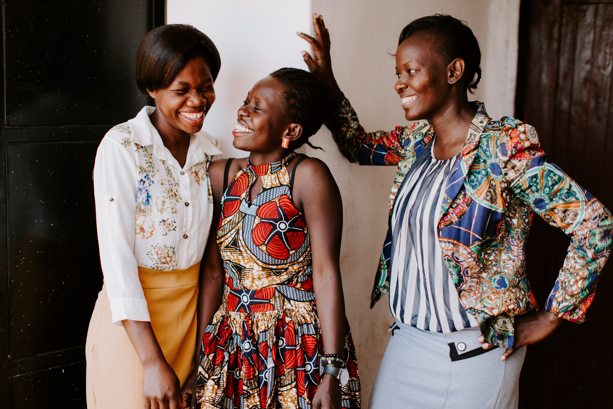 Three women stand, two lean into each other sharing a joke, while the other laughs too.