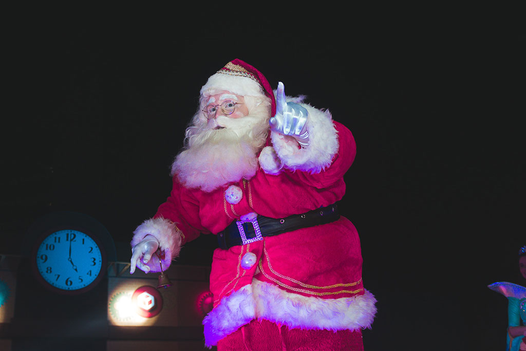 A Santa tiptoes in a darkened room carrying a lamp and holding up a finger in a gesture of silencing