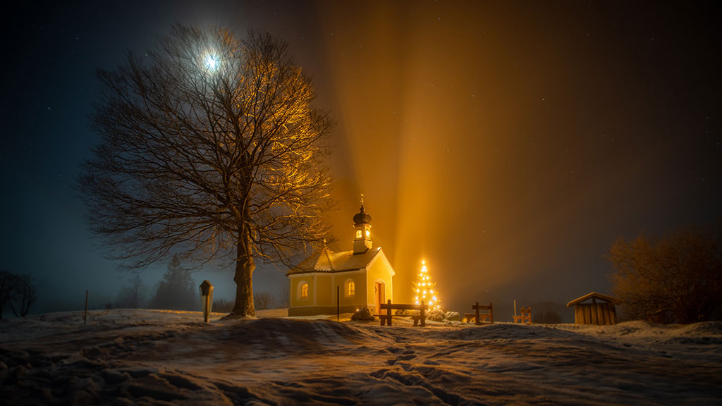 A nocturnal snow-covered scene of a tree, chapel and Christmas tree casting shadows.