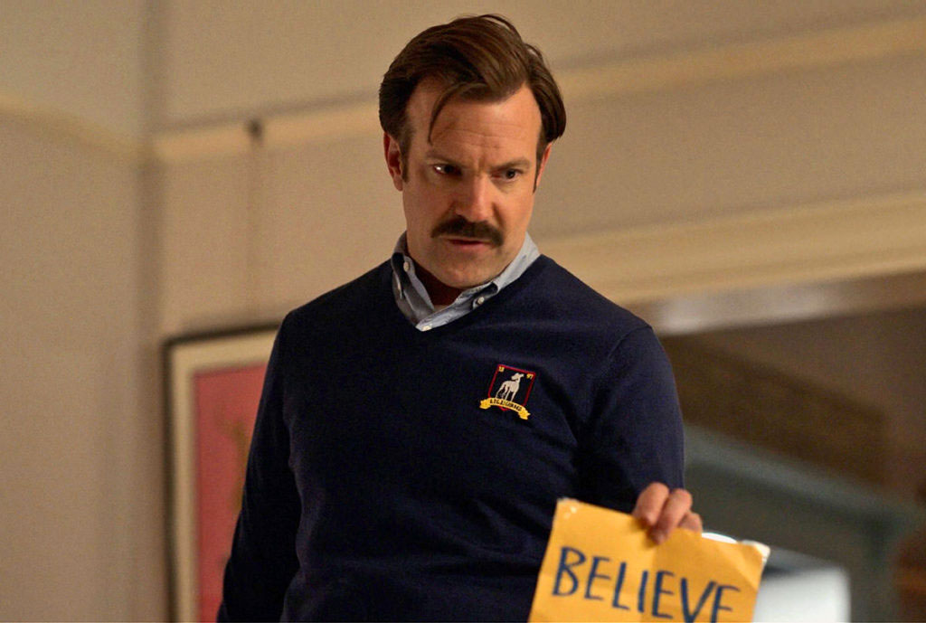 A man in a blue jumper holds a yellow sign reading 'believe'.