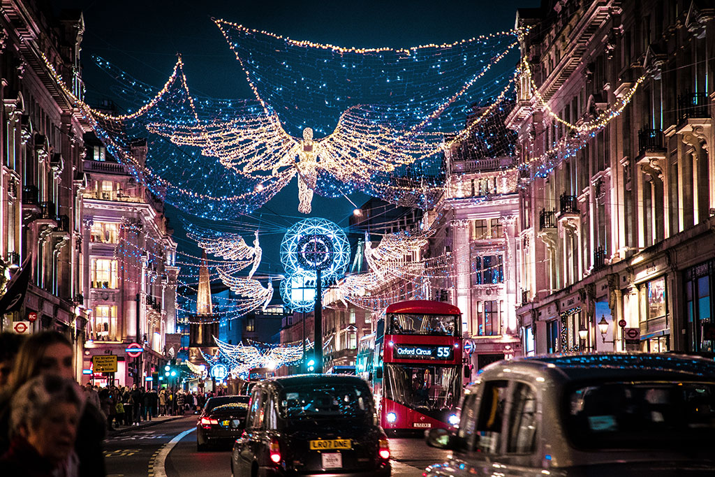 A shopping street is crowded by taxi cabs and buses while above it a Christmas illumination of an angel hangs over all.