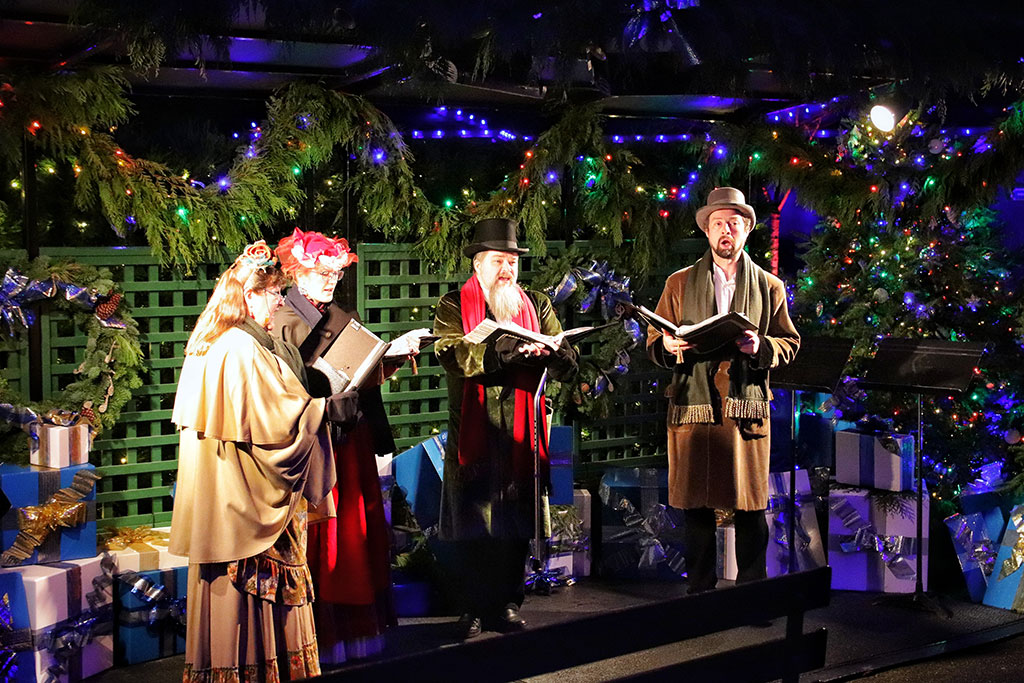 Dressed in Victorian clothes, a group of carol singers stand and sing amid Christmas foliage.