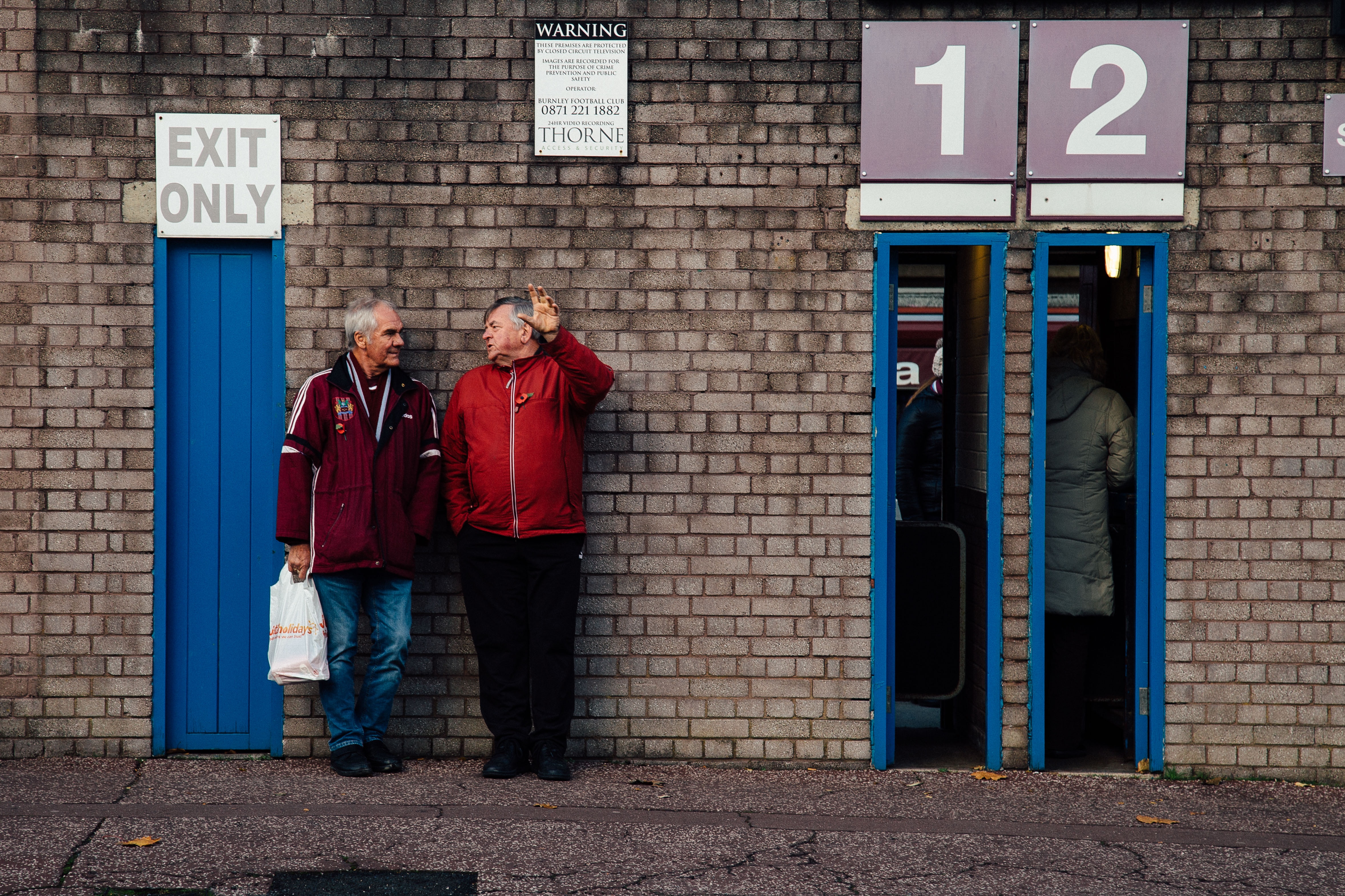 Two older men lean agains the wall of a football club's entrance, next to thin open doorways.